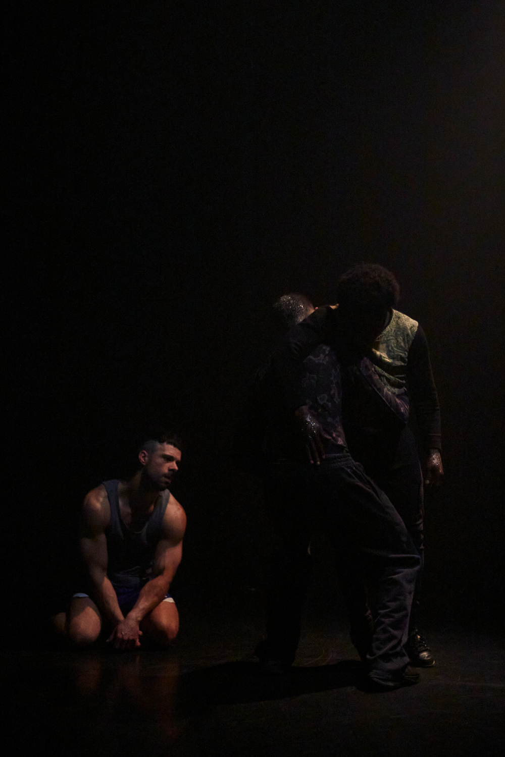 Deux danseurs sont enlacés et un danceur est à genou et les observe, création lumière de Jérôme Baudouin, créateur lumière en danse, théâtre et spectacle vivant